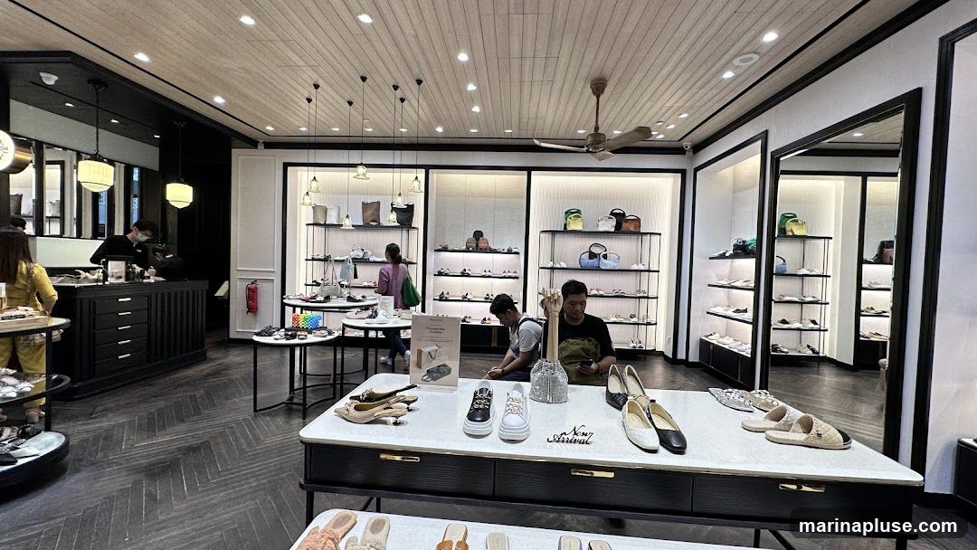 The interior of a well-lit shoe store with displays of shoes and handbags on shelves and tables. Customers are browsing, and staff are attending to them. The store has a modern and clean aesthetic with wooden floors and black and white accents.