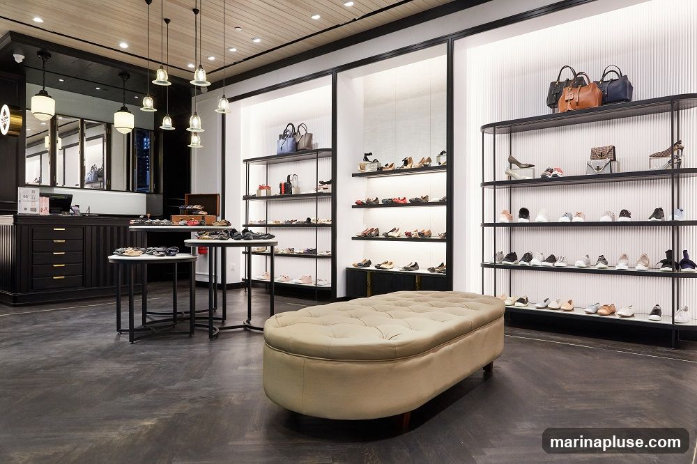 A well-lit shoe store interior with displays of women's shoes and handbags. The store features dark wood flooring, black shelving units, and a large tufted ottoman in the center. Shoes are neatly arranged on shelves, and handbags are displayed on the top shelves of some units. A counter with drawers is visible on the left side.