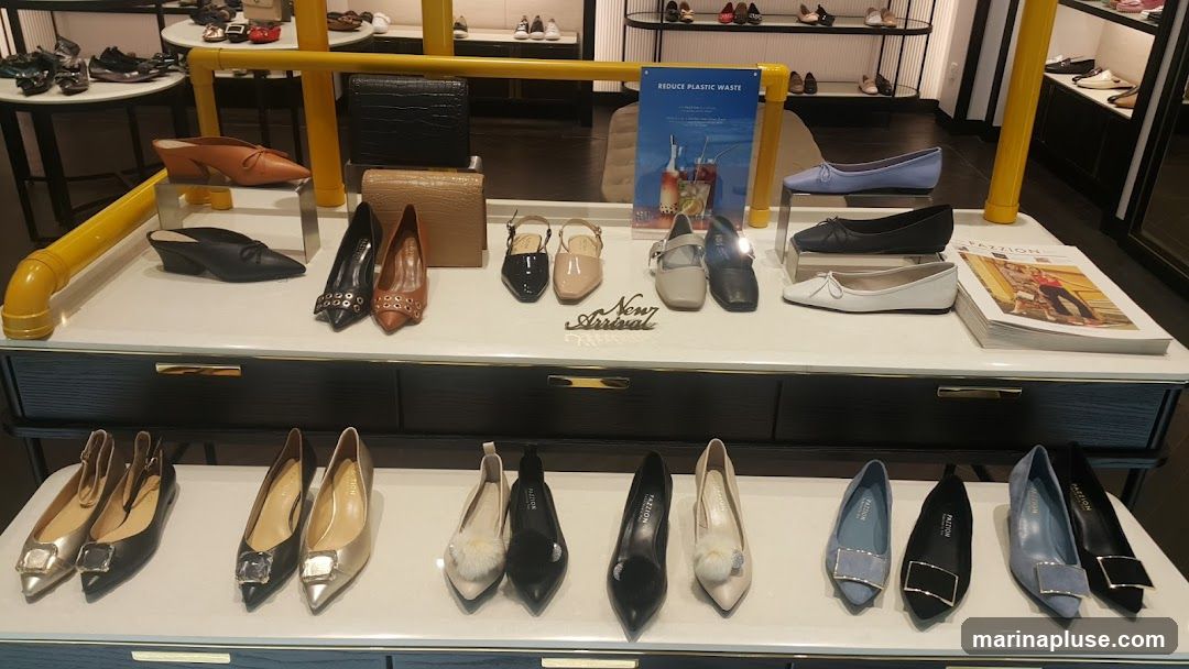 A display of various women's shoes, including flats, heels, and loafers, arranged on white shelves and a black cabinet. Some shoes have decorative buckles or fur accents. A magazine titled "PAZZION" is also visible. A sign promoting plastic waste reduction is displayed in the background.