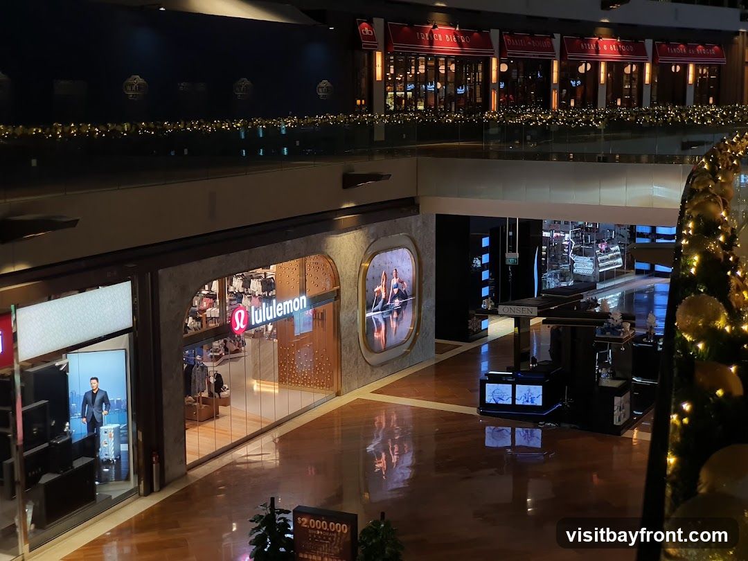 A view of a Lululemon store entrance within a shopping mall. The store features its logo and displays clothing inside. Adjacent to it, another storefront shows a digital advertisement. Further into the mall, other shops and displays are visible, including a Christmas tree decorated with lights and ornaments. The mall has polished wooden floors and is decorated with festive lights along the railings.