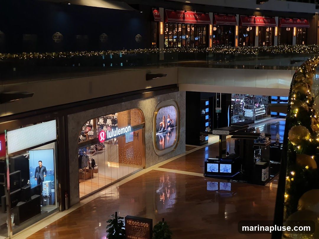 A view of a Lululemon store entrance within a shopping mall. The store features its logo and displays clothing inside. Adjacent to it, another storefront shows a digital advertisement. Further into the mall, other shops and displays are visible, including a Christmas tree decorated with lights and ornaments. The mall has polished wooden floors and is decorated with festive lights along the railings.