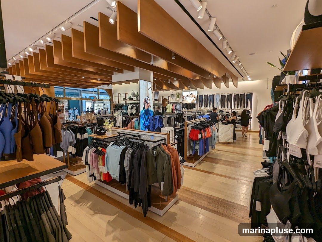 A wide shot of the interior of a retail store selling athletic apparel. Racks of clothing, including sports bras and leggings, are displayed on wooden floors. The store features a modern design with wooden slatted ceilings and track lighting. Several customers are browsing the merchandise, and staff members are visible near a checkout counter. A large poster featuring a model in athletic wear is displayed on a wall.