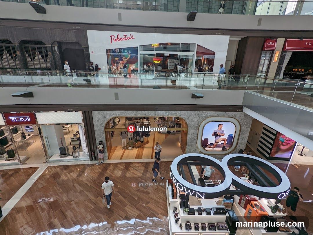 An overhead view of a shopping mall interior. On the upper level, a restaurant with the name 'Roberto's' is visible. On the lower level, the storefronts of Tumi and Lululemon are prominent. A kiosk with the word 'CROWN' is also visible, displaying watches and other accessories. Several shoppers are seen walking around the mall.