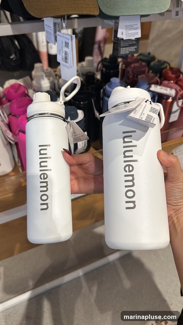 Two white Lululemon water bottles are displayed on a shelf. The bottles have the Lululemon logo printed vertically on them. In the background, there are more water bottles in various colors and hats on a shelf above.