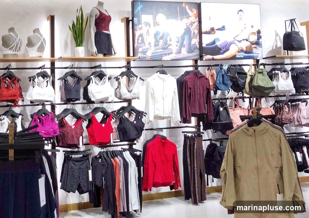 A retail display featuring athletic wear, including sports bras, jackets, leggings, and bags. Mannequins showcase some of the apparel. Digital screens on the wall display people exercising.