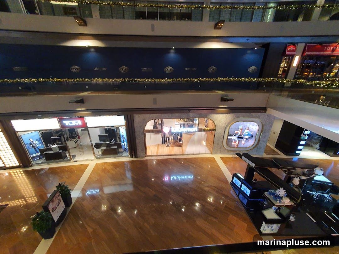 An interior view of a multi-level shopping mall. In the foreground, a polished wooden floor reflects the overhead lights. To the left, a store with a prominent "TUMI" sign displays merchandise in its windows. Adjacent to it, another store with a rounded entrance features the "lululemon" logo and displays mannequins. A large digital screen is embedded in the wall next to the lululemon store, showing an advertisement. Further back, a dark blue wall is adorned with repeating circular logos and festive gold garlands. Other shop fronts are visible on different levels, including one with a red sign that appears to say "FRENCH." A black display stand with various items is positioned in the lower right corner.
