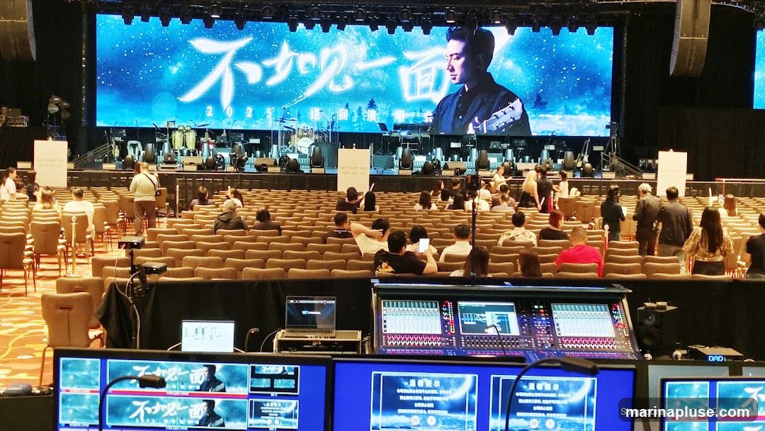 A large screen displays a performer and Chinese text, with musical instruments set up on stage. Rows of empty seats face the stage, and a sound mixing board and monitors are visible in the foreground.