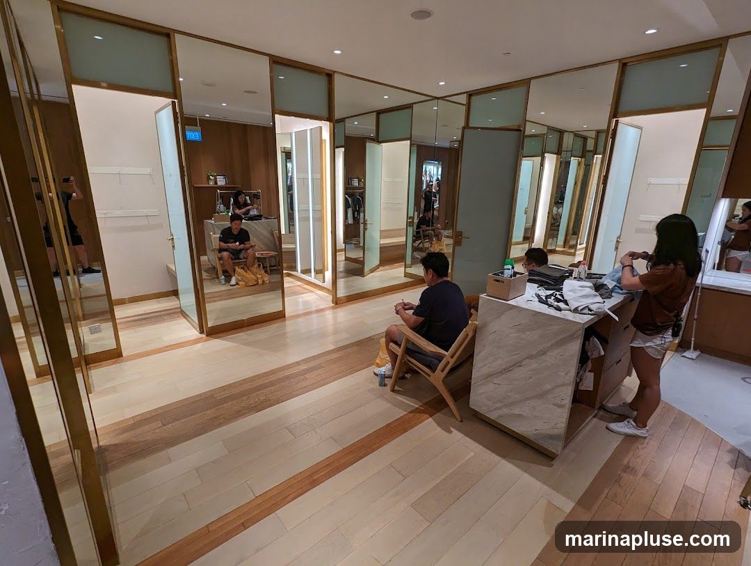 The image shows the interior of a clothing store with multiple fitting rooms. The fitting rooms have mirrored doors and are decorated with wood paneling and brass accents. Several people are visible in the image, including customers waiting and staff members attending to them. There is a counter with clothing items and a chair for customers to sit on.