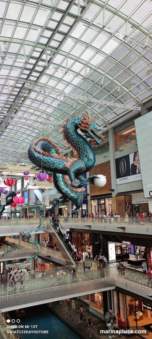 An interior view of a multi-level shopping mall, featuring a large, ornate dragon sculpture suspended from the ceiling. The mall has a glass roof, escalators, and numerous shops with illuminated displays. People are seen walking on the various levels and escalators. Decorative elements like paper lanterns and string lights are also visible. Brands like Chanel and Kate Spade are identifiable on storefronts.