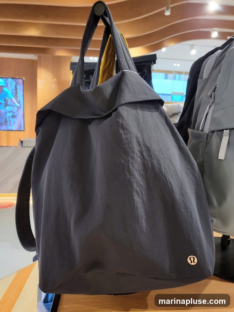 A dark gray tote bag with a yellow scarf draped inside is displayed on a hook. The bag has a subtle sheen and a logo on the lower right corner. In the background, other bags and a screen displaying an image are visible.