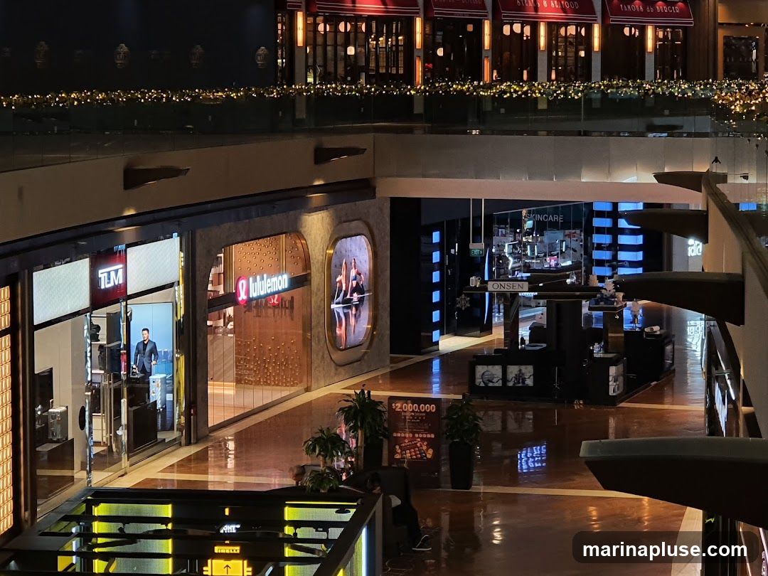 An interior view of a shopping mall with multiple storefronts visible. On the left, a store with a sign that reads "TLM" is seen, with a man standing inside. Next to it is a "Lululemon" store. Further down, a store labeled "SKINCARE" and "ONSEN" is visible. The mall has a polished floor reflecting the lights and decorations.