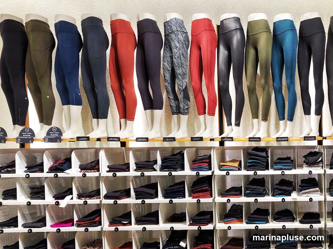 A wall display of various leggings on mannequins and folded in cubbies. The leggings are in a variety of colors and patterns, including black, olive green, navy blue, red, grey, and a black and white pattern. Some of the leggings have visible brand tags. Below the mannequins, folded leggings are neatly organized in white cubbies.