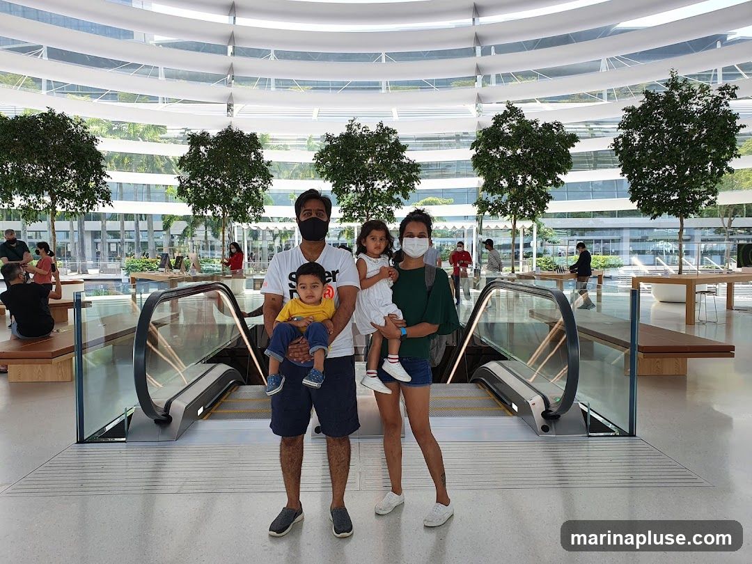 A family of four, two adults and two young children, pose for a photo in a spacious, modern store with a high, curved ceiling and large windows. The store features escalators, trees planted indoors, and display tables with electronics. The family is wearing masks, and the children are being held by the adults.