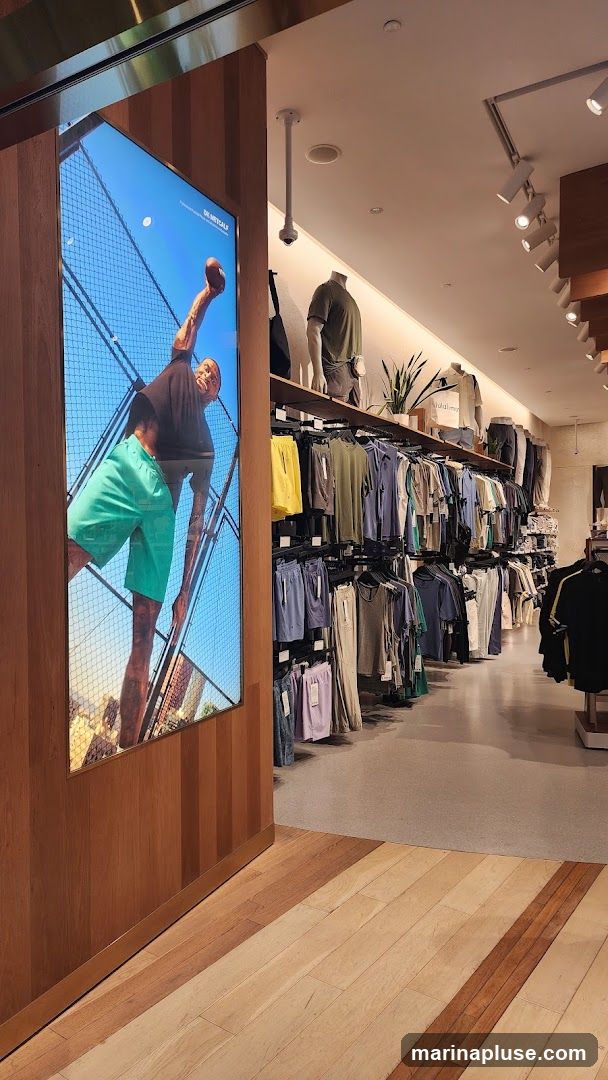 A well-lit clothing store interior with racks of athletic wear. A large digital display on the left shows a man in athletic shorts and a t-shirt. Racks of t-shirts and shorts in various colors are displayed on the right. Mannequins are also visible.