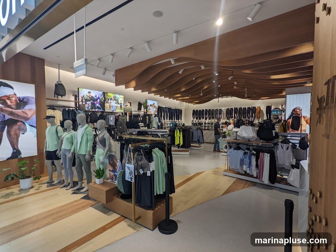 The interior of a brightly lit athletic apparel store. Mannequins dressed in athletic wear are displayed at the front of the store. Racks of clothing, including leggings, tops, and outerwear, are visible throughout the space. Large screens display images of people engaged in athletic activities. The store features a modern design with wooden accents on the ceiling and walls.
