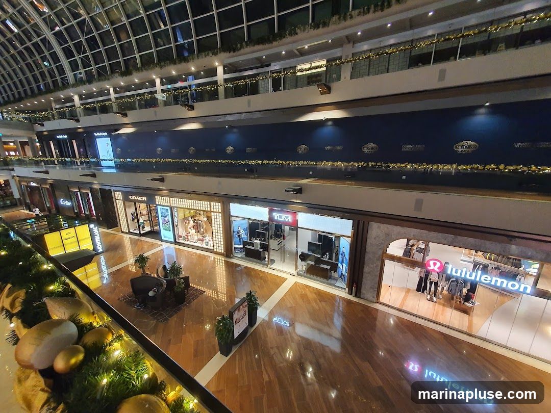 An interior view of a multi-level shopping mall with various storefronts visible. The mall features a glass ceiling, wooden floors, and decorative garlands with lights. Stores like Cartier, Coach, Tumi, and Lululemon are identifiable. A "Coming Soon" banner is displayed on an upper level.
