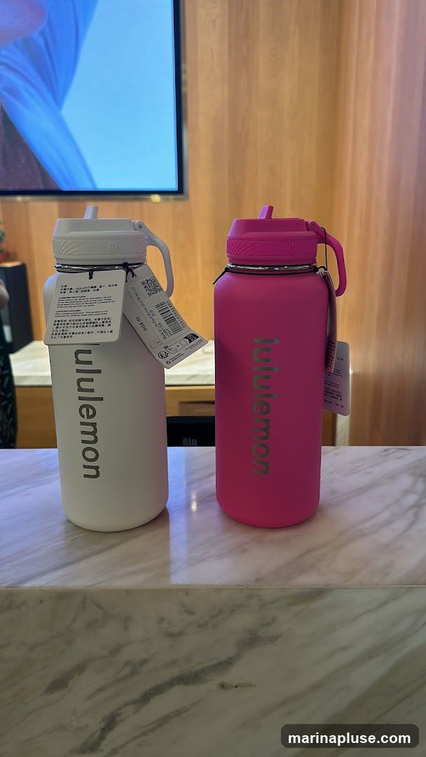 Two Lululemon water bottles, one white and one pink, are displayed on a marble counter. Both bottles have the Lululemon logo and name printed vertically. The white bottle has a tag with product information and a price of $39.95. In the background, a screen displays an image and a wooden wall is visible.