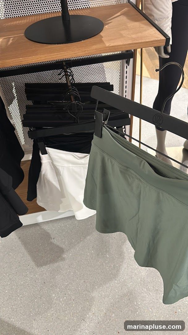 A close-up view of athletic skirts displayed on hangers in a retail store. The skirts are in white and a muted green color. A mannequin wearing black leggings is visible in the background. The store's branding, a circular logo, is visible on a hanger.
