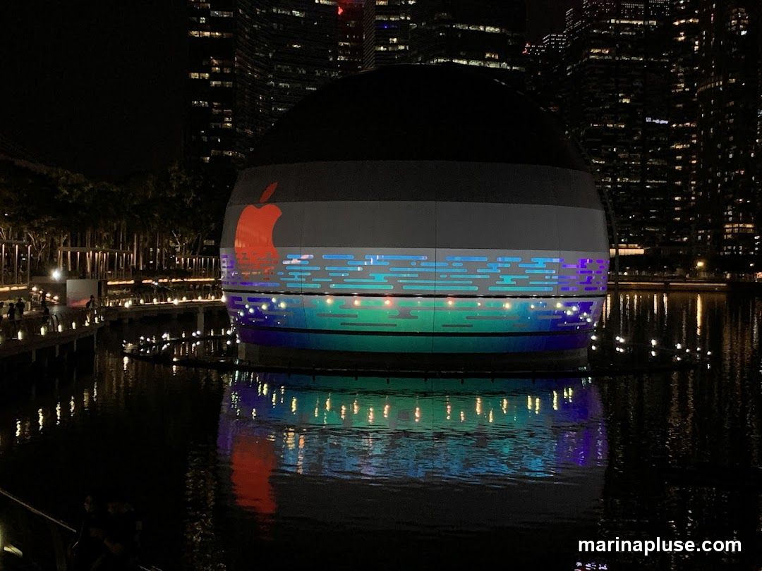 A spherical Apple Store building at night, illuminated with colorful lights and featuring the iconic Apple logo. The building is situated on the water, with reflections visible on the surface. The city skyline is visible in the background.
