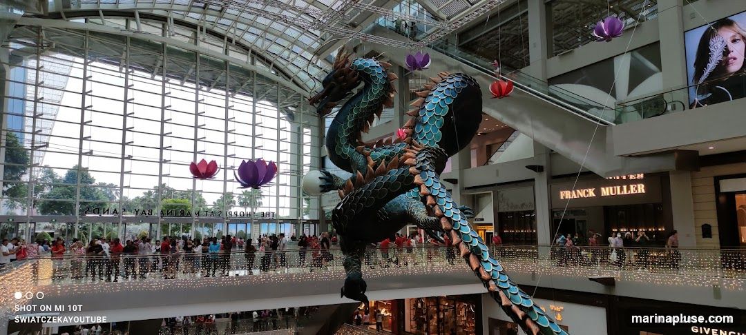 A large, ornate dragon sculpture hangs from the ceiling of a modern shopping mall. The dragon is predominantly teal and blue with orange accents. It is surrounded by decorative lotus flowers in pink and purple. Below, people are gathered on balconies and walkways, looking up at the sculpture. The mall features large glass windows, escalators, and storefronts, including visible signage for "FRANCK MULLER" and "GIVENCHY". The overall scene suggests a festive or decorative display within a busy commercial space.