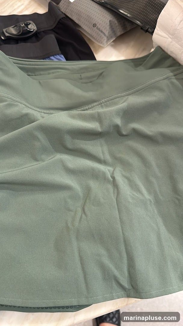 A close-up view of a green athletic skirt. The fabric appears to be lightweight and slightly textured. The waistband is visible at the top, and the hem of the skirt is at the bottom. In the background, other clothing items and a Lululemon branded pouch are visible.