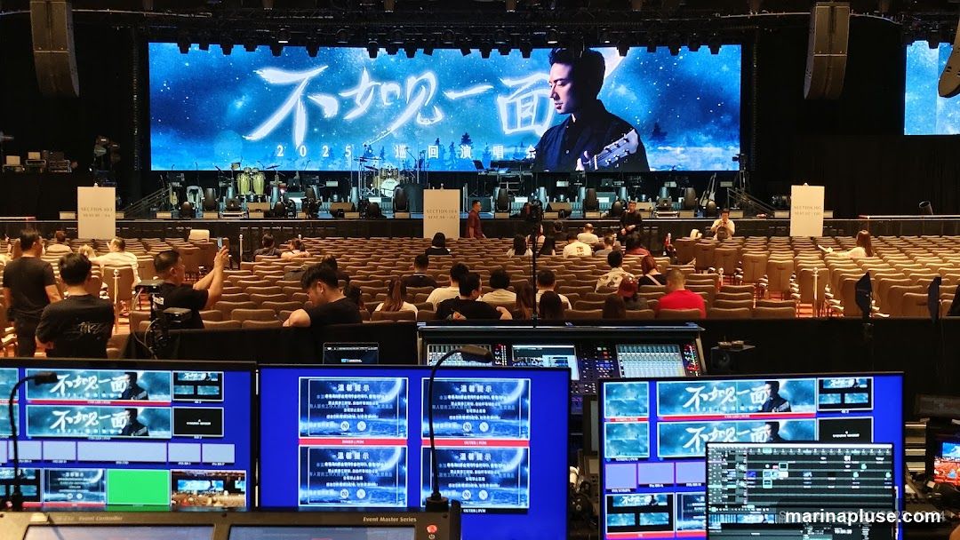 A wide shot of a concert venue, showing a large stage with a performer on a screen, musical instruments, and rows of empty seats. In the foreground, a control room is visible with multiple monitors displaying stage feeds and production controls. The date and location 'Singapore - 12/20/2025, 19:04' are visible on one of the monitors.