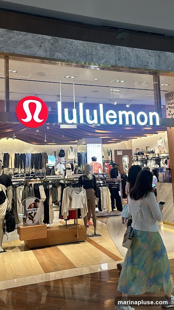 The exterior of a Lululemon store is visible, with the brand's logo and name prominently displayed above the entrance. Racks of athletic apparel are displayed inside, with mannequins showcasing outfits. Several customers are seen browsing the merchandise.