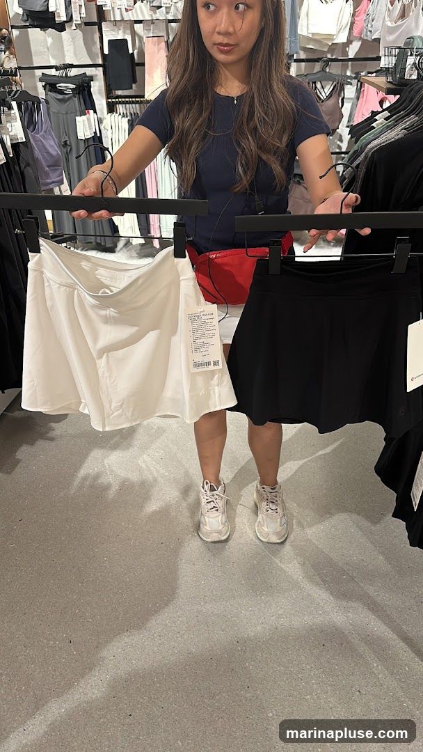 A woman stands in a store, holding up two skirts on hangers. The skirt on the left is white, and the skirt on the right is black. She is wearing a navy blue t-shirt and a red fanny pack. The background shows racks of clothing and store fixtures.