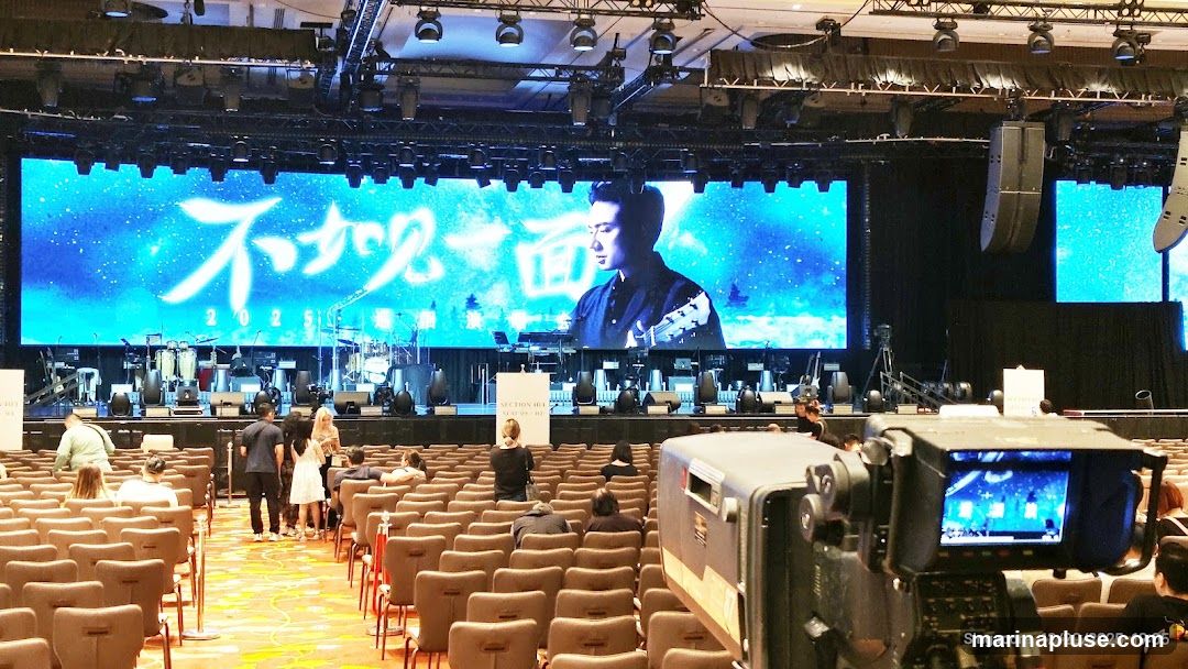 A wide shot of a concert stage with a large LED screen displaying a man playing a guitar and Chinese characters. The screen also shows the year 2025. The stage is equipped with musical instruments and lighting rigs. In front of the stage, rows of empty seats are set up, with a few people milling about. A camera is positioned in the foreground, facing the stage. The overall scene suggests a pre-event setup for a concert or performance.