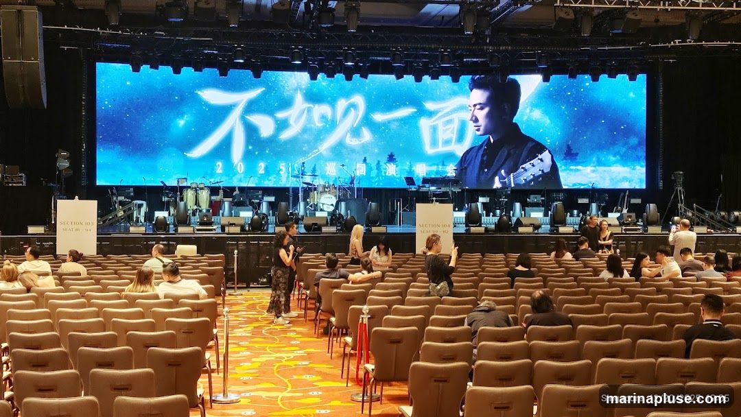 A wide shot of a concert stage with a large LED screen displaying an image of a man playing a guitar and Chinese characters. The stage is set up with musical instruments, including drums and keyboards. In front of the stage, rows of empty seats are visible, with a few audience members scattered throughout. The lighting is dim, with spotlights focused on the stage. The date and location "Singapore 12/20/2025, 19:05" are visible in the bottom right corner.