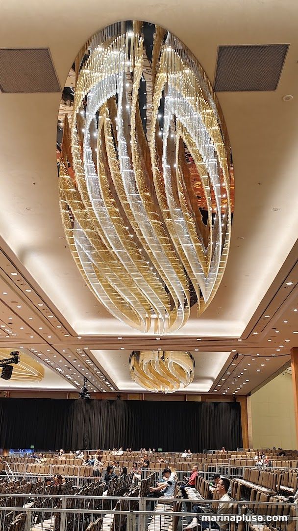 A large, ornate chandelier hangs from the ceiling of a spacious ballroom. The chandelier is oval-shaped and features multiple tiers of gold and silver elements that reflect light. Below, rows of empty seats are arranged in a theater-like setting, with a black curtain backdrop. Several people are seated in the rows, facing a stage area that is not fully visible. The overall impression is of a venue prepared for an event.