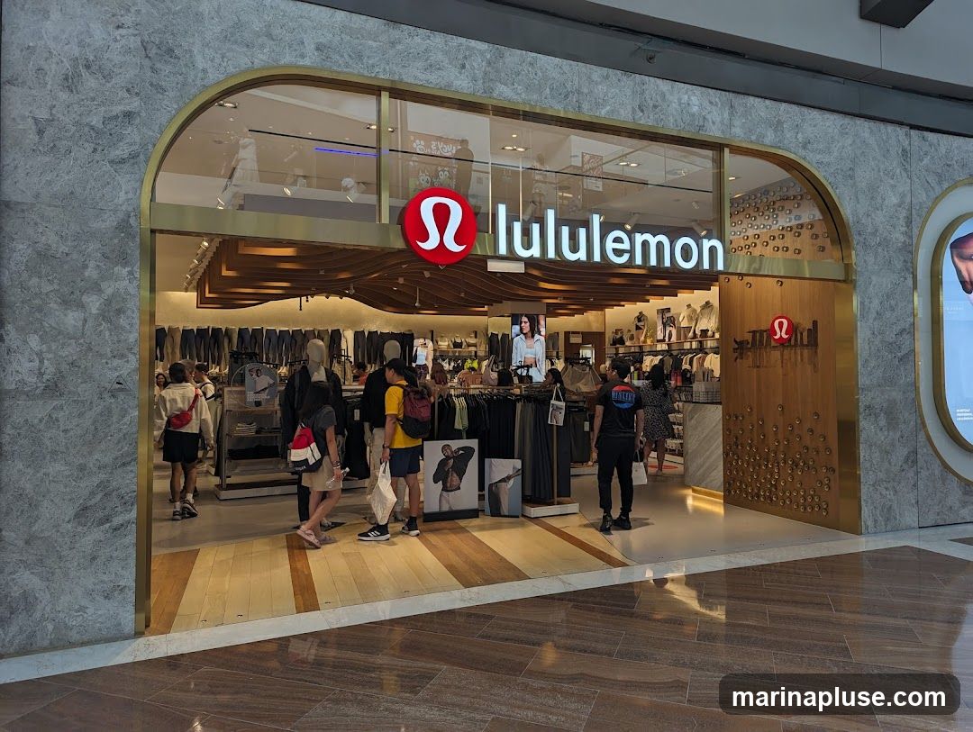 The exterior of a Lululemon store with its prominent logo and name displayed above the entrance. People are seen browsing inside the store, which is filled with athletic apparel. The store features a modern design with wooden accents and marble-like walls.