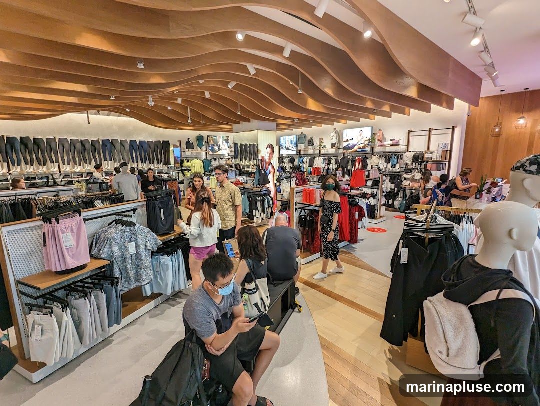 A wide shot of the interior of a brightly lit athletic apparel store. The store features a modern design with a wavy wooden ceiling. Racks of clothing, including leggings, shorts, and shirts, are displayed throughout the store. Several customers are browsing the merchandise, and some staff members are visible. Mannequins are also present, showcasing outfits. The overall atmosphere is clean and organized, typical of a retail environment.