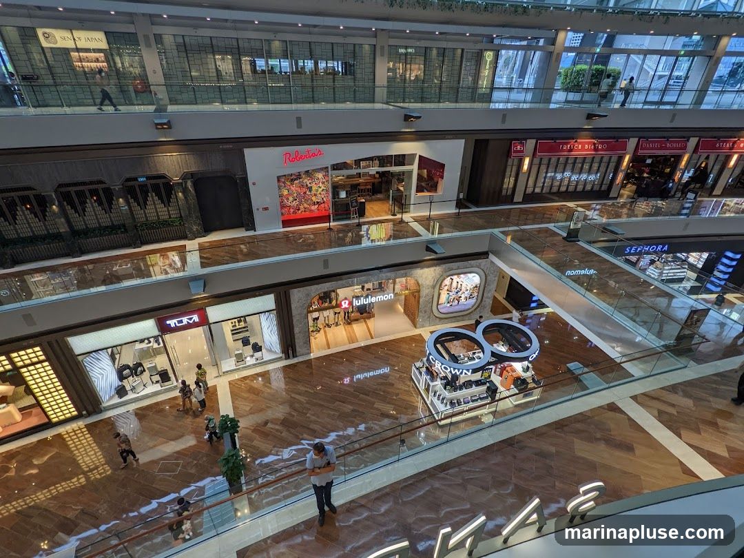 An interior view of a modern shopping mall with multiple levels. Shops such as TUMI, lululemon, Sephora, and various restaurants are visible. People are seen walking around and browsing the stores. The mall features polished wooden floors and glass railings.