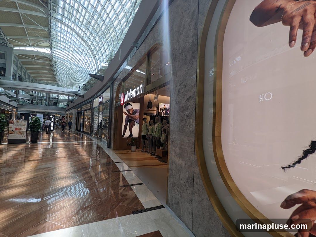 A view of a Lululemon store entrance in a modern shopping mall. The store features a large display window with mannequins dressed in athletic wear. The Lululemon logo is prominently displayed above the entrance. The mall has a distinctive arched glass ceiling that allows natural light to illuminate the polished floor. Other shops and architectural details of the mall are visible in the background.