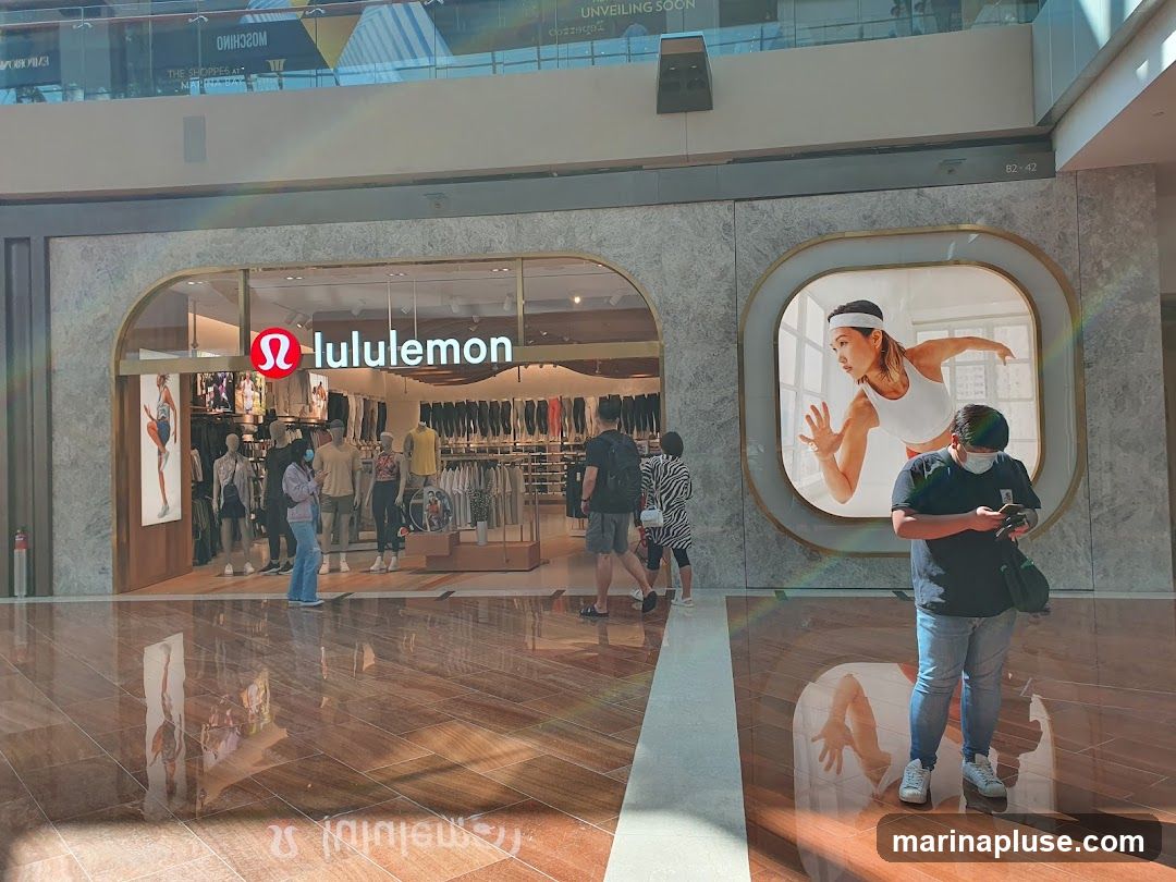 The exterior of a Lululemon store with its logo and name prominently displayed above the entrance. Inside, mannequins showcase athletic wear, and racks of clothing are visible. A large digital display featuring a woman running is mounted on the wall to the right of the entrance. Several people are seen entering and browsing the store, while one person stands outside looking at their phone.
