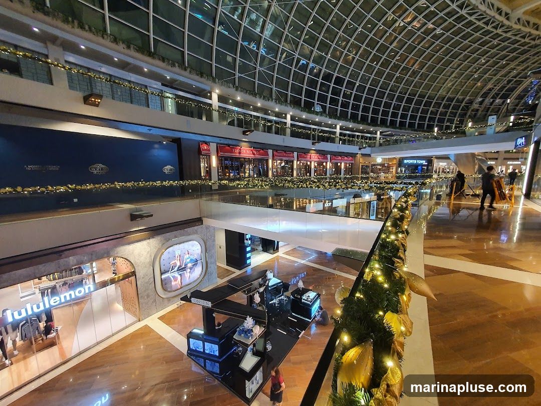 An interior view of a multi-level shopping mall, adorned with festive gold and green garlands and lights. Various shops are visible on different levels, including Lululemon and Sports Town. A central atrium area features a black display stand with products. People are seen walking and working within the mall. The ceiling is a distinctive glass and metal grid structure.