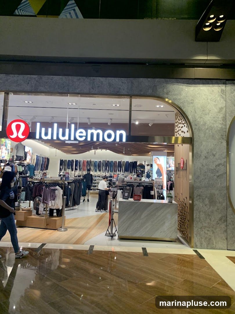 The exterior of a Lululemon store is visible, with the brand's logo and name prominently displayed above the entrance. Inside, racks of athletic apparel are displayed, and customers and staff can be seen browsing and interacting. The store features a modern interior design with marble accents and warm lighting.