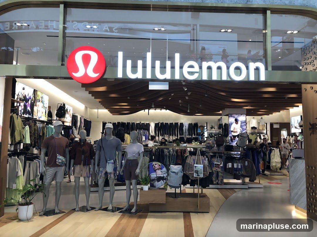 The exterior of a Lululemon store with its prominent logo and name displayed above the entrance. Inside, mannequins are dressed in athletic wear, and racks of clothing are visible. The store appears to be located in a shopping mall, with other shops and shoppers visible in the background through the glass facade.