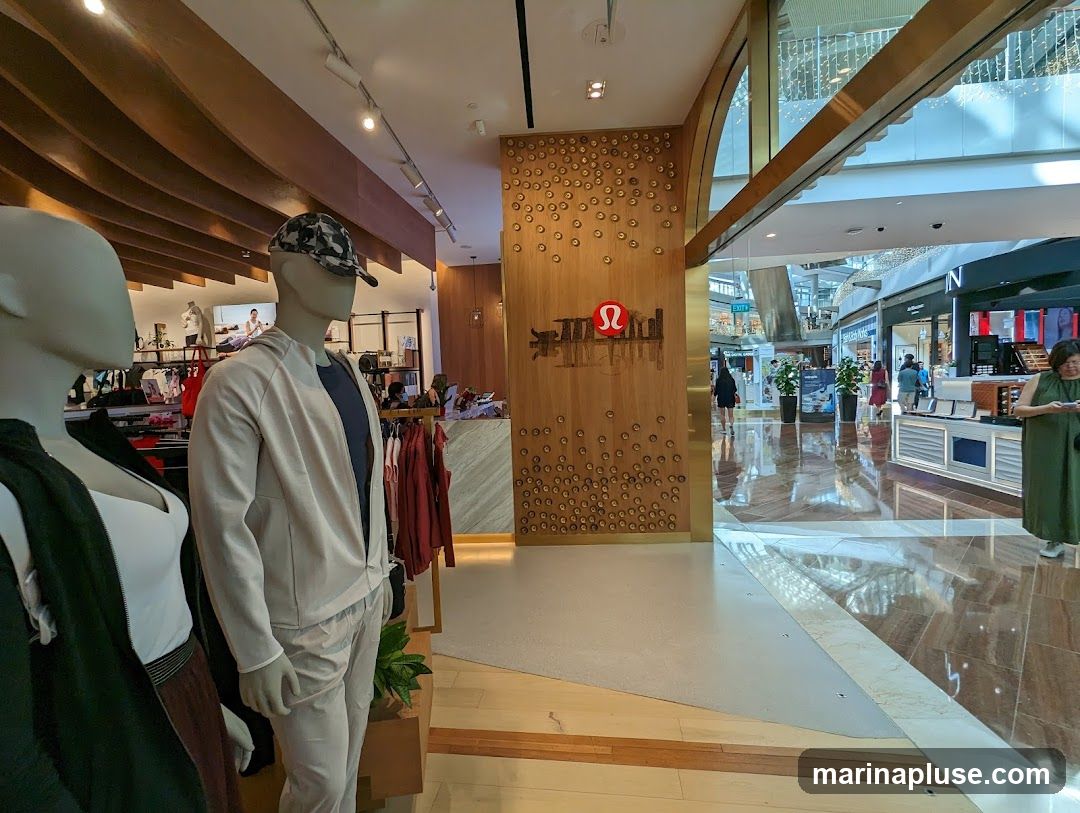 Mannequins dressed in athletic wear are displayed inside a Lululemon store. The store features a wooden accent wall with the Lululemon logo and a cityscape design. Racks of clothing are visible, along with other shoppers and store displays in the background.