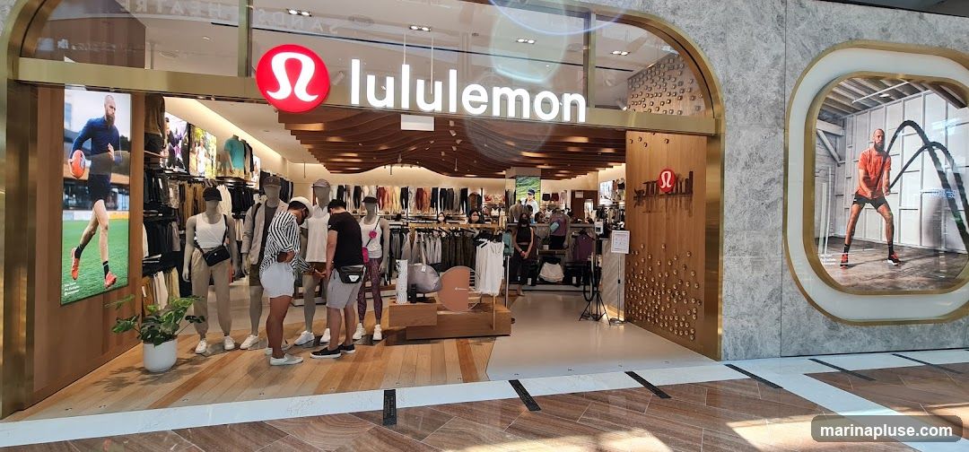 The exterior of a Lululemon store. The store features a large sign with the Lululemon logo and name above the entrance. Inside, clothing is displayed on racks and mannequins. Large digital screens on either side of the entrance show athletic imagery.