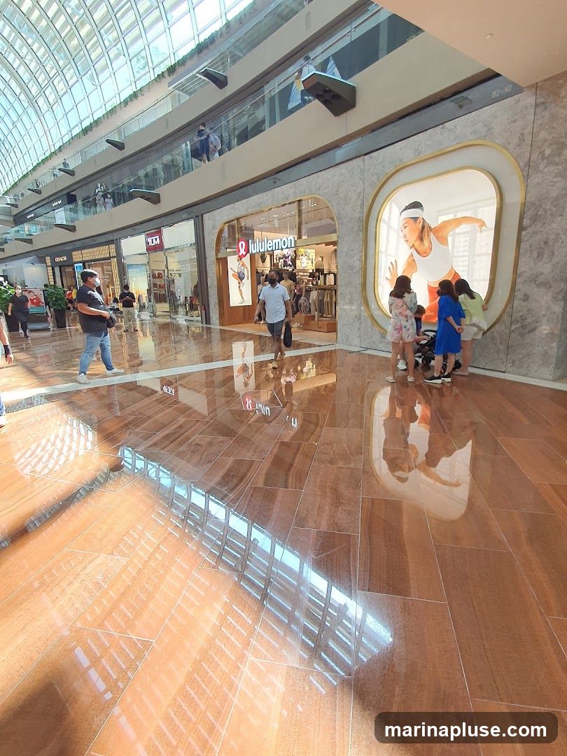 The exterior of a Lululemon store is visible, with its logo and name displayed above the entrance. Inside, clothing racks and displays can be seen. To the right of the entrance, a large illuminated display features an image of a woman in athletic wear. Several people are gathered in front of the display. The store is located within a modern shopping mall with a glass ceiling and multiple levels.