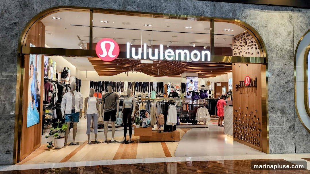 The exterior of a Lululemon store with mannequins displaying athletic wear. The store features a large sign with the Lululemon logo and name. Inside, racks of clothing and accessories are visible.