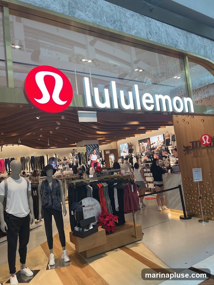 The exterior of a Lululemon store with its prominent red and white logo and "lululemon" text signage. Mannequins dressed in athletic wear are displayed outside the entrance, along with racks of clothing. The store interior is visible, showing more apparel and customers.