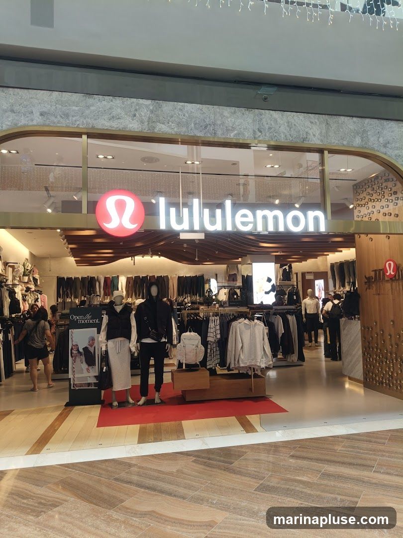 The exterior of a Lululemon store is visible, featuring the brand's logo and name prominently displayed above the entrance. Inside, mannequins are dressed in athletic wear, and racks of clothing are displayed. The store appears to be located within a shopping mall, with other shops and shoppers visible in the background.