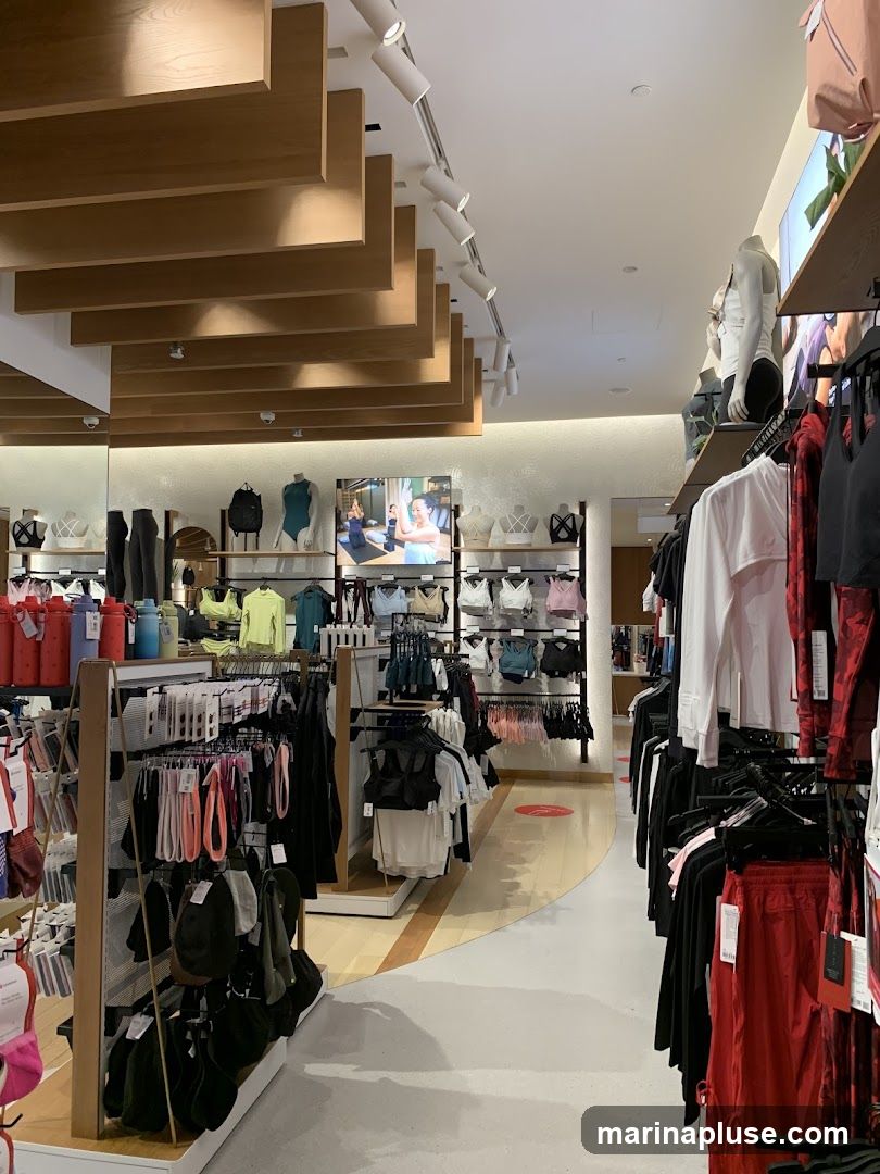 The interior of a brightly lit athletic apparel store. Racks of clothing, including sports bras, leggings, tops, and shorts, are displayed on shelves and hanging racks. Mannequins are also present, showcasing outfits. A large screen on the back wall displays a video of people exercising. The store features a modern design with wooden accents on the ceiling and shelves.