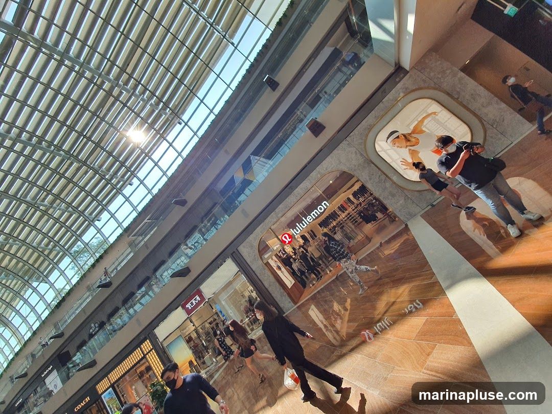 A view inside a Lululemon store with various apparel displayed. People are walking around the store. The store has a modern interior design with a large display featuring athletic figures. The ceiling of the mall is visible, with sunlight streaming through.