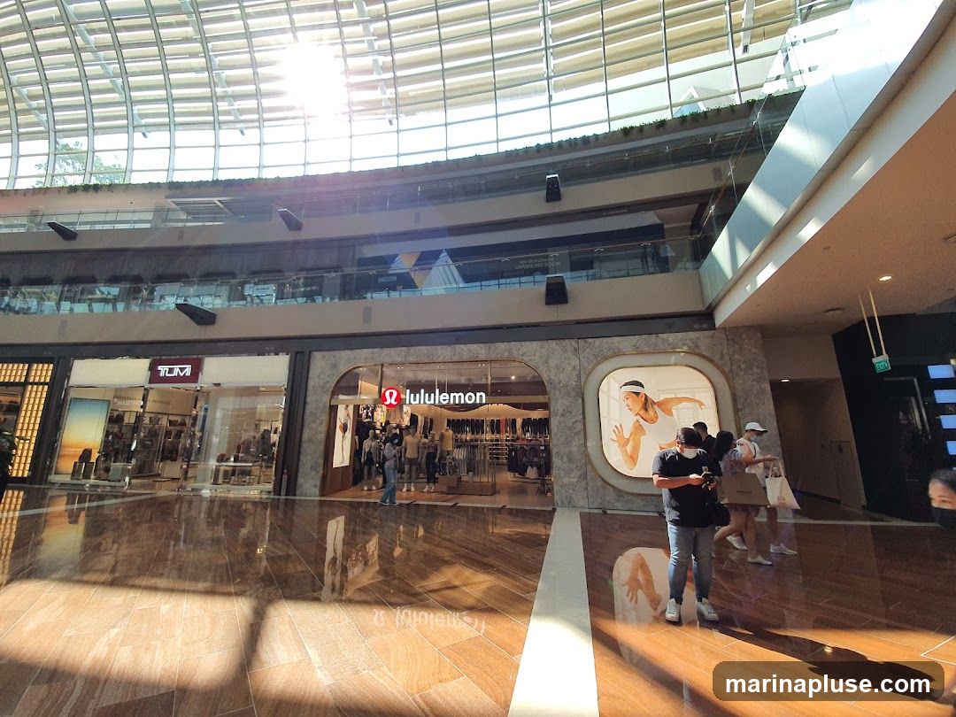 The storefront of a Lululemon store is visible, featuring the brand's logo and an interior display of athletic apparel. Adjacent to it is a Tumi store. The stores are located within a modern shopping mall with a large, arched glass ceiling that allows natural light to illuminate the polished wooden floor.