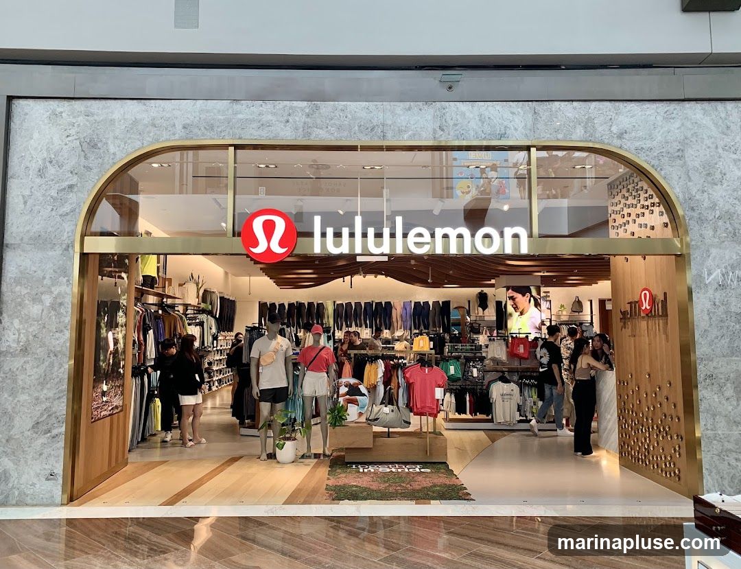The exterior of a Lululemon store with its prominent logo and name displayed above the entrance. The store is well-lit and showcases athletic apparel on racks and mannequins. Several customers are visible inside the store.