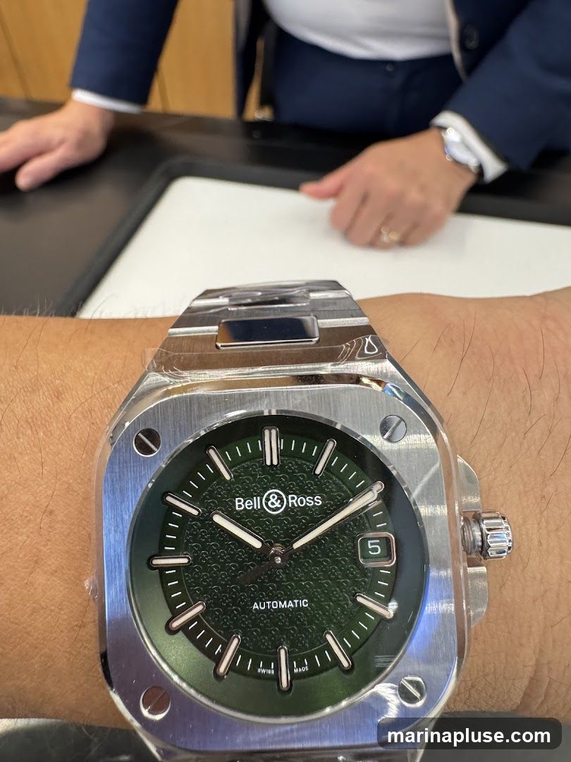 A close-up shot of a Bell & Ross automatic watch with a green dial, worn on a person's wrist. The watch features a square stainless steel case and a date window displaying the number 5. In the background, two individuals are partially visible, appearing to be in a retail or service environment.